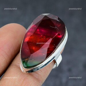 Könnte beinhalten: Ein großer, tropfenförmiger Ring mit einem facettierten, mehrfarbigen Edelstein. Der Edelstein wechselt von tiefrot zu grün und ist in einem silbernen Band gefasst. Der Ring wird am Finger getragen.