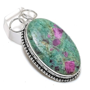 Könnte beinhalten: Ovaler Anhänger mit einem grünen Stein mit leuchtend rosa Einschlüssen, gefasst in einem silberfarbenen Metallrahmen. Der Anhänger hat einen dekorativen, perlenbesetzten Rand und eine Öse mit drei Ringen.