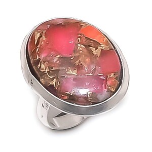 Könnte beinhalten: Ein ovaler Ring mit einer polierten, silberfarbenen Metallschiene und Fassung. Das Herzstück ist ein glatter, mehrfarbiger Steineinsatz mit leuchtend pinken, dunkelroten und metallisch kupferfarbenen Sprenkeln auf der gesamten Oberfläche.