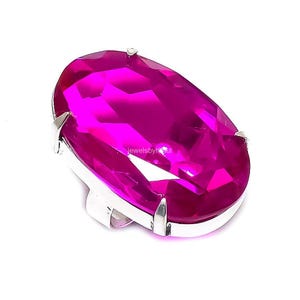 Könnte beinhalten: Ein Ring mit einem großen, oval geschliffenen, leuchtend pinkfarbenen Edelstein in einer polierten, silberfarbenen Krappenfassung aus Metall. Der facettierte Stein reflektiert das Licht auf seiner Oberfläche, und die Ringschiene ist unter der Fassung sichtbar.