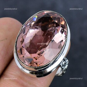Könnte beinhalten: Silberner Ring mit einem großen, oval geschliffenen rosa Edelstein. Der Edelstein ist facettiert und reflektiert das Licht, wodurch verschiedene Rosatöne entstehen. Der Ring hat ein Blumenmuster an der Seite.