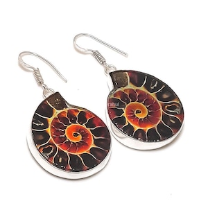 Puede incluir: Un par de pendientes colgantes con fósiles de amonita pulidos engastados en metal plateado. Cada pendiente muestra un patrón de concha en espiral con tonos marrón oscuro, negro y naranja vibrante, suspendidos de ganchos plateados simples.