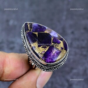 Könnte beinhalten: Ein tropfenförmiger Ring mit einem großen, polierten violett-goldenen Kupfer-Türkis-Edelstein, gefasst in ein dekoratives Sterlingsilberband mit detaillierten Perlen- und Filigranverzierungen entlang der Ränder der Metallfassung.