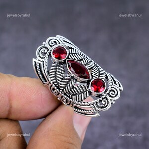 Op de afbeelding: Een zilverkleurige ring met drie rode edelstenen en een bladerenontwerp. De ring heeft een ovale vorm met een centrale marquise-geslepen steen en twee ronde stenen. De ring wordt door een hand vastgehouden.