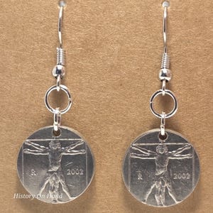 May include: A pair of silver-toned dangle earrings. Each earring features a circular pendant with the Vitruvian Man design and the year 2002. The earrings have a simple hook and a small bead detail.