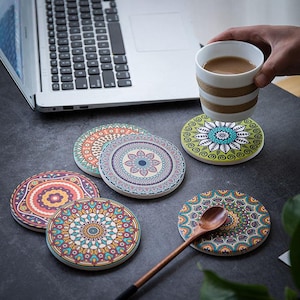 May include: A collection of colorful ceramic coasters with intricate mandala designs. The coasters feature various patterns in shades of blue, green, orange, and purple. A wooden spoon and a cup of coffee are also present, suggesting a home decor theme.
