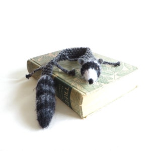 May include: A handmade crochet raccoon bookmark in shades of gray and white, lying on top of a vintage book with green and white floral patterns. The raccoon has a long tail and outstretched arms.