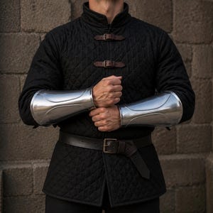 May include: A black quilted long-sleeve tunic featuring a high collar and two brown leather buckle straps across the chest. The garment is paired with polished silver metal forearm bracers and a brown leather belt with a metal buckle at the waist.