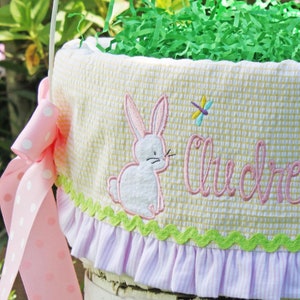 May include: An Easter basket with a white handle and a cream-colored fabric lining. The fabric is embroidered with two bunnies and the name "Audrey Rose" in pink. The basket is filled with green paper grass and two orange carrot decorations. Pink ribbons adorn the sides.