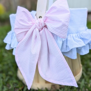 May include: A decorative basket with a large pink and white striped bow. The basket has a light blue and white striped fabric trim with a ruffled edge. The basket is sitting on green grass.