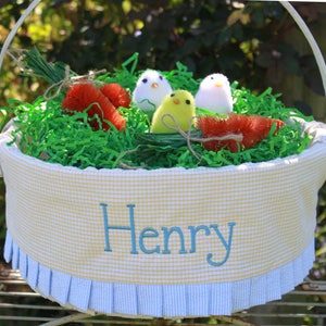 May include: A round Easter basket with a yellow and white gingham pattern, blue trim, and the name "Henry" embroidered in blue. The basket is filled with green paper grass, toy chicks, and carrot decorations.