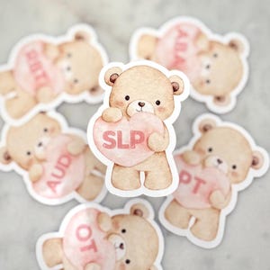 Peut inclure: Plusieurs autocollants représentant des ours en peluche à l'aquarelle. Chaque ours tient un cœur rose avec du texte, dont "SLP", "AUD", "OT", "PT" et "ABA". Les ours sont brun clair avec des yeux et un nez noirs.