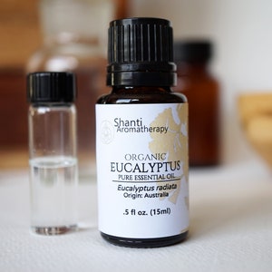 May include: A small brown glass bottle with a black cap containing organic eucalyptus essential oil. The label is white with black text and reads "Shanti Aromatherapy, Organic Eucalyptus, Pure Essential Oil, Eucalyptus radiata, Origin: Australia, .5 fl oz. (15ml)"