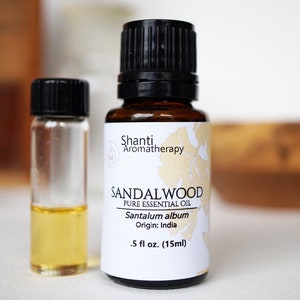 Op de afbeelding: Een bruine glazen fles met een zwarte dop en een wit etiket met de tekst "Shanti Aromatherapy Sandalwood Pure Essential Oil Santalum album Origin: India 0,5 fl oz. (15ml)"