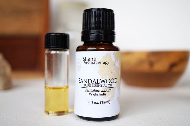 Op de afbeelding: Een bruine glazen fles met een zwarte dop en een wit etiket met de tekst "Shanti Aromatherapy Sandalwood Pure Essential Oil Santalum album Origin: India 0,5 fl oz. (15ml)"
