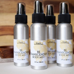 Puede incluir: Cuatro botellas de spray plateadas con tapas negras y etiquetas blancas. Las etiquetas dicen "Shanti Aromatherapy" y "Be Transcendent Body Mist" con un diseño floral. Cada botella contiene 90 ml de bruma corporal.
