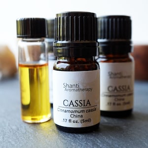 May include: Three small brown glass bottles with black caps containing essential oils. The center bottle is labeled "Shanti Aromatherapy Cassia Cinnamomum cassia China .17 fl oz. (5ml)"