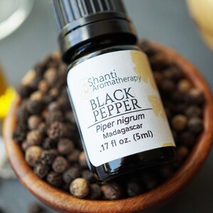 May include: A close-up of a bottle of Shanti Aromatherapy Black Pepper essential oil. The bottle is .17 fl oz (5ml) and sits in a wooden bowl filled with black peppercorns. The label reads "Piper nigrum Madagascar".