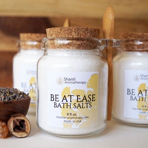 May include: Three glass jars with cork lids and wooden spoons. The jars contain white bath salts and have labels that read "Shanti Aromatherapy" and "Be At Ease Bath Salts". The jars are on a wooden surface.