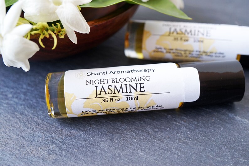 May include: A white and gold label on a clear glass roller bottle with a black cap. The label reads "Shanti Aromatherapy Night Blooming Jasmine .35 fl oz 10ml". The bottle is sitting on a gray surface.