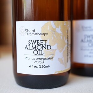 Puede incluir: Dos botellas de vidrio &aacute;mbar de aceite de almendras dulces. Las etiquetas dicen "Shanti Aromatherapy" y "Sweet Almond Oil, Prunus amygdalus dulcis, 120ml". El fondo est&aacute; borroso.