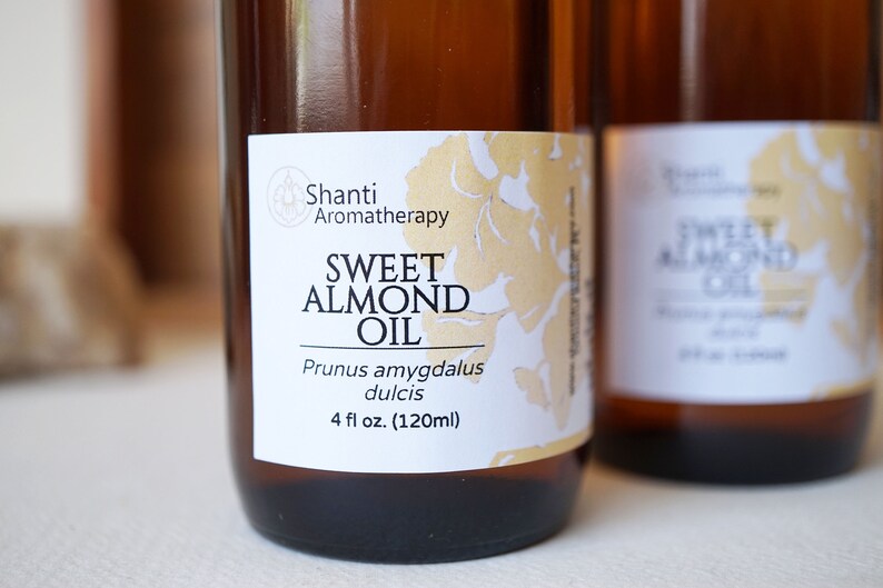Puede incluir: Dos botellas de vidrio &aacute;mbar de aceite de almendras dulces. Las etiquetas dicen "Shanti Aromatherapy" y "Sweet Almond Oil, Prunus amygdalus dulcis, 120ml". El fondo est&aacute; borroso.