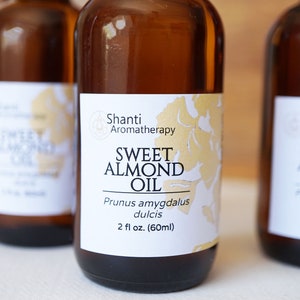 Puede incluir: Tres botellas de vidrio marr&oacute;n de Shanti Aromatherapy Sweet Almond Oil. Cada botella tiene una etiqueta blanca con detalles florales dorados y el texto "Sweet Almond Oil" y "2 fl oz. (60ml)". Las botellas est&aacute;n dispuestas en fila.