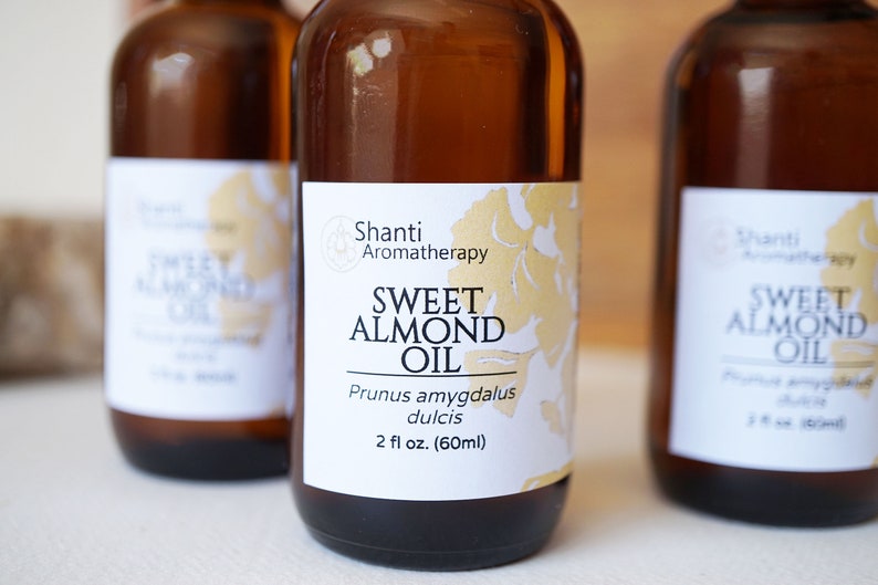 Puede incluir: Tres botellas de vidrio marr&oacute;n de Shanti Aromatherapy Sweet Almond Oil. Cada botella tiene una etiqueta blanca con detalles florales dorados y el texto "Sweet Almond Oil" y "2 fl oz. (60ml)". Las botellas est&aacute;n dispuestas en fila.