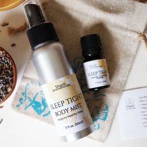 May include: A silver spray bottle with a black cap and a white label that reads "Shanti Aromatherapy Sleep Tight Body Mist Supports Healthy Sleep 3 fl oz (90ml)". A small brown glass bottle with a black cap and a white label that reads "Shanti Aromatherapy Sleep Tight Lavender Chamomile Roman Chamomile 1/2 fl oz (15ml)". A small white bag with a brown drawstring. A brown card with the text "thank you" written in black ink. A small bowl filled with dried lavender flowers.