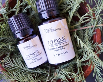 Op de afbeelding: Twee amberkleurige glazen flesjes met essentiële cypres-olie, gelabeld met "Shanti Aromatherapy" en "CYPRESS PURE ESSENTIAL OIL", met de botanische naam en herkomst van de olie, "Cupressus sempervirens Herkomst: Kreta", en het volume, "5 fl oz. (15ml)" en "17 fl oz. (5ml)", respectievelijk. De flesjes liggen in een houten kom gevuld met groen blad.