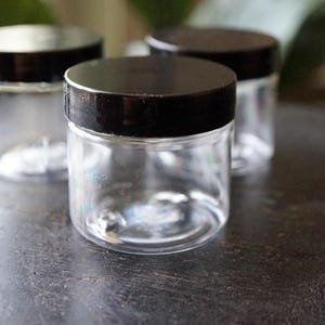 May include: Three clear, small glass jars with black lids are arranged on a dark, textured surface. The jars are cylindrical with a wide opening and a rounded base. The lids are solid black and screw-on style. The jars are empty.