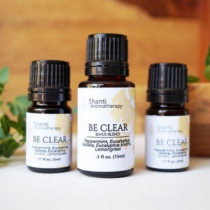 May include: Three brown glass bottles with black caps and white labels. The labels read "Shanti Aromatherapy" and "BE CLEAR SINUS BLEND". The bottles contain essential oil blends for aromatherapy. The bottles are on a white surface with a wooden bowl and green leaves in the background.