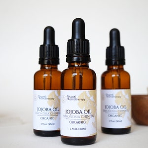 Puede incluir: Tres botellas de vidrio ámbar con goteros negros llenas de aceite de jojoba. Las botellas están etiquetadas con "Jojoba Oil Simmondsia Chinensis Organic 1 fl oz. (30ml)" y "Shanti Aromatherapy".