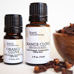 May include: Two amber glass bottles with black caps containing orange clove essential oil blend. The bottles are labeled "Shanti Aromatherapy" and "Orange Clove Essential Oil Blend". The labels list the ingredients as "Blood Orange, Frankincense, Clove Bud". The bottles are sitting on a white surface with dried cloves scattered around them.