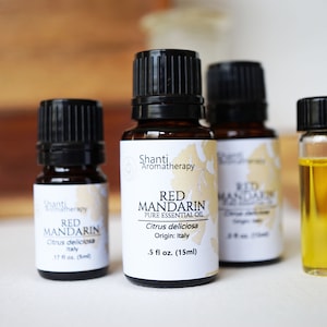 Puede incluir: Tres botellas de vidrio ámbar con tapas negras que contienen aceite esencial de mandarina roja. Las botellas están etiquetadas con "Shanti Aromatherapy" y "Red Mandarin Pure Essential Oil". La etiqueta también indica "Citrus deliciosa Origin: Italy" y el tamaño es de 0,5 fl oz (15 ml).
