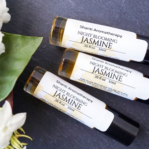 May include: Three 10 ml roll-on bottles of Shanti Aromatherapy Night Blooming Jasmine essential oil. The bottles are on a dark gray surface with white jasmine flowers in the background.