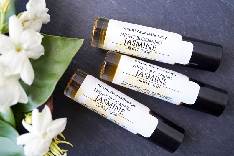 May include: Three 10 ml roll-on bottles of Shanti Aromatherapy Night Blooming Jasmine essential oil. The bottles are on a dark gray surface with white jasmine flowers in the background.