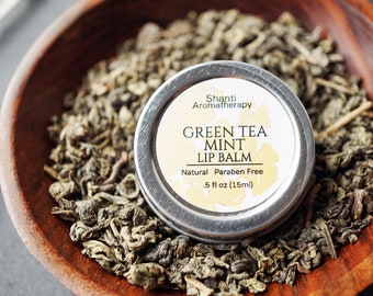 May include: A silver tin of Shanti Aromatherapy Green Tea Mint Lip Balm. The tin is sitting on a bed of loose green tea leaves. The label reads "Shanti Aromatherapy Green Tea Mint Lip Balm, Natural Paraben Free, .5 fl oz (15ml)"