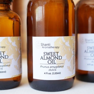 Puede incluir: Varias botellas de vidrio &aacute;mbar de aceite de almendras dulces. Cada botella tiene una etiqueta blanca con detalles florales dorados y el texto "Sweet Almond Oil" y "Shanti Aromatherapy". Las botellas son de 60 ml y 120 ml.