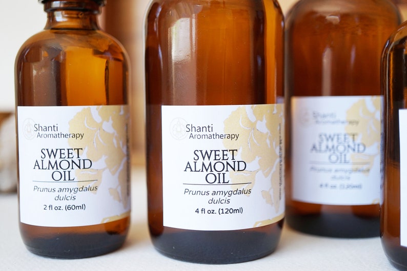 Puede incluir: Varias botellas de vidrio &aacute;mbar de aceite de almendras dulces. Cada botella tiene una etiqueta blanca con detalles florales dorados y el texto "Sweet Almond Oil" y "Shanti Aromatherapy". Las botellas son de 60 ml y 120 ml.