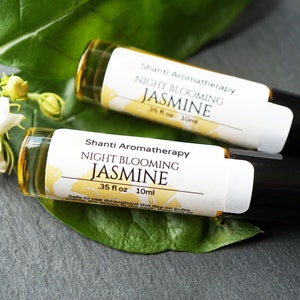 May include: Two small glass bottles with white labels that read "Shanti Aromatherapy Night Blooming Jasmine .35 fl oz 10ml". The bottles are on a gray surface with a green leaf.