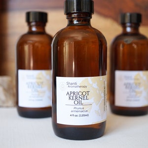 May include: Three amber glass bottles with black caps and white labels. The center bottle label reads "Shanti Aromatherapy Apricot Kernel Oil Prunus armeniaca 4 fl oz (120ml)"