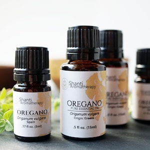 May include: Four amber glass bottles of oregano essential oil with black caps. The bottles are labeled with the brand name "Shanti Aromatherapy" and the words "Oregano Pure Essential Oil". The bottles are on a black surface with a sprig of oregano in the background.