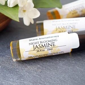 May include: A small glass bottle with a black rollerball top and a white label that reads "Shanti Aromatherapy Night Blooming Jasmine .35 fl oz 10ml". The bottle is filled with a yellow liquid and is sitting on a gray surface. The label also includes the text "Safe to use throughout the day on pulse points, the wrists, the neck. Keep out of direct sunlight and heat."