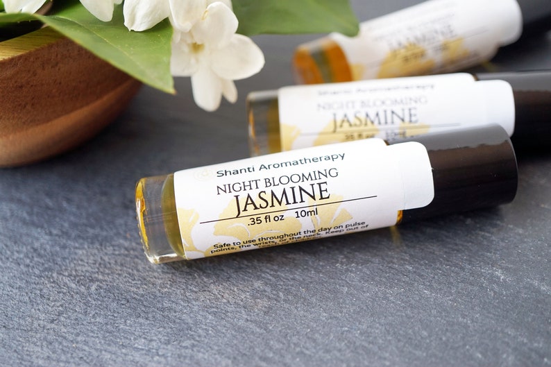 May include: A small glass bottle with a black rollerball top and a white label that reads "Shanti Aromatherapy Night Blooming Jasmine .35 fl oz 10ml". The bottle is filled with a yellow liquid and is sitting on a gray surface. The label also includes the text "Safe to use throughout the day on pulse points, the wrists, the neck. Keep out of direct sunlight and heat."
