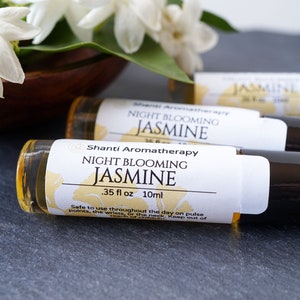 May include: Three small glass bottles with black roll-on tops and white labels. The labels read "Shanti Aromatherapy Night Blooming Jasmine .35 fl oz 10ml". The bottles are on a gray surface with white jasmine flowers in the background.