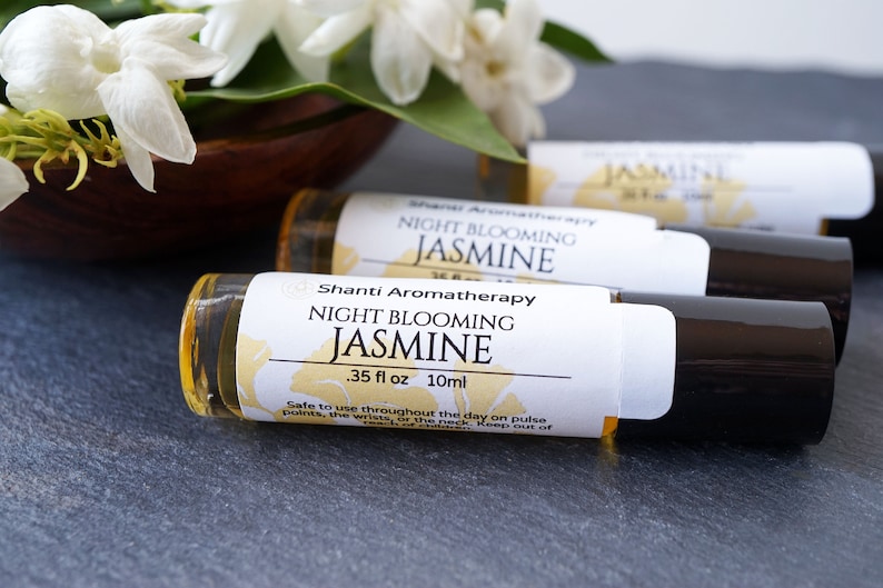 May include: Three small glass bottles with black roll-on tops and white labels. The labels read "Shanti Aromatherapy Night Blooming Jasmine .35 fl oz 10ml". The bottles are on a gray surface with white jasmine flowers in the background.