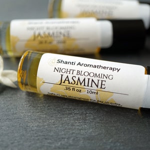 May include: A small amber glass bottle with a white label that reads "Shanti Aromatherapy Night Blooming Jasmine .35 fl oz 10ml Safe to use throughout the day on pulse points". The bottle is lying on a gray surface with a white jasmine flower in the foreground.