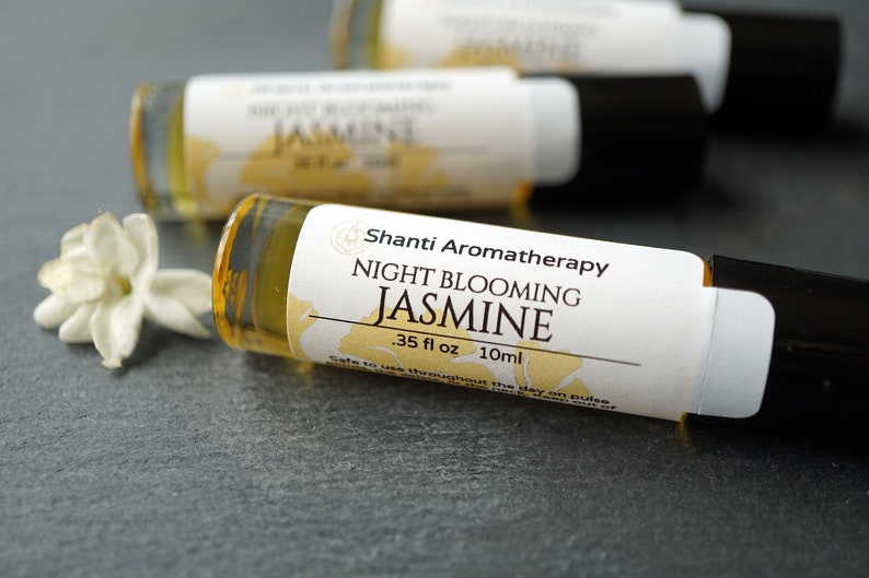 May include: A small amber glass bottle with a white label that reads "Shanti Aromatherapy Night Blooming Jasmine .35 fl oz 10ml Safe to use throughout the day on pulse points". The bottle is lying on a gray surface with a white jasmine flower in the foreground.