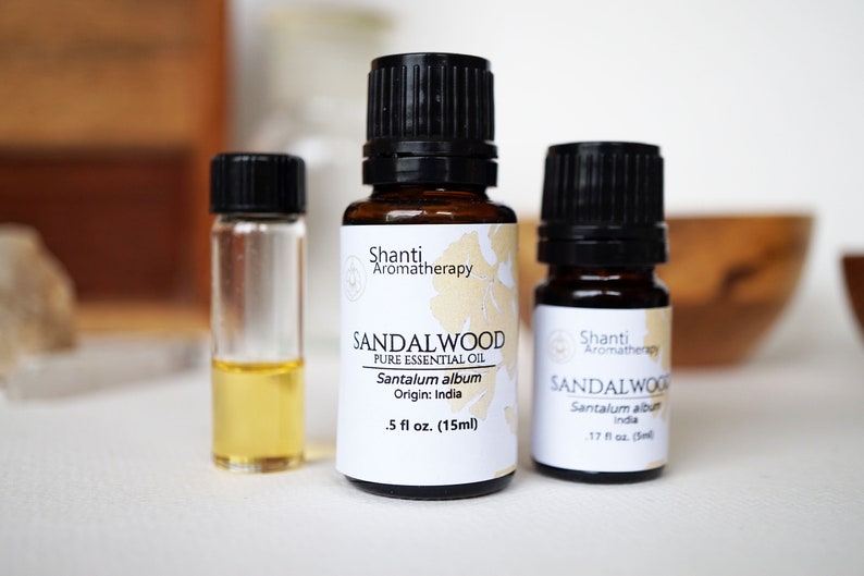 Op de afbeelding: Twee bruine glazen flesjes met sandelhout etherische olie met zwarte dopjes en witte etiketten. De etiketten lezen "Shanti Aromatherapy" en "Sandalwood Pure Essential Oil Santalum album Origin: India". Het kleinere flesje is 0,5 fl oz (15 ml) en het grotere flesje is 0,17 fl oz (5 ml).